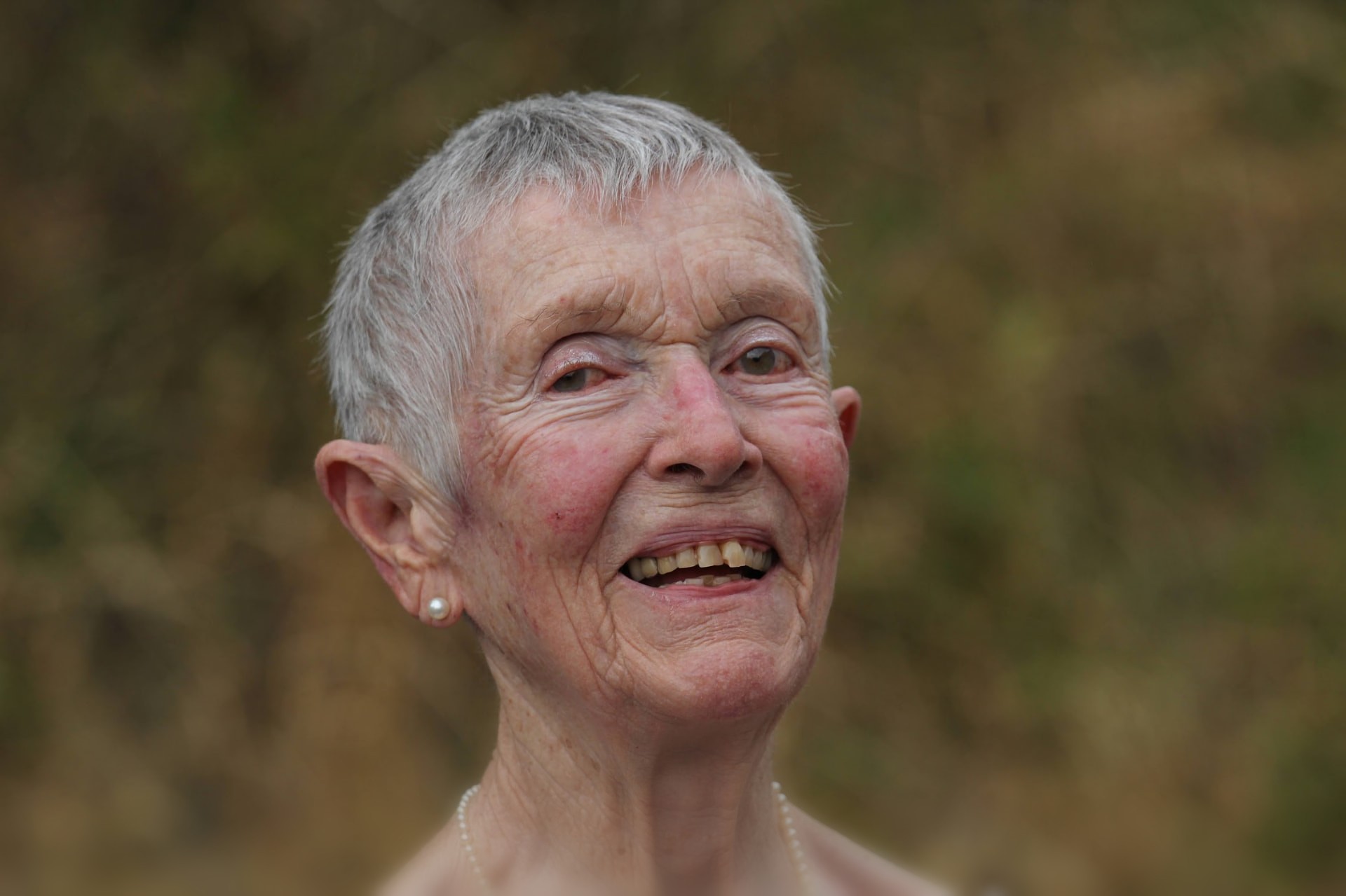 Femme âgée aux cheveux gris et collier de perles, souriante en extérieur